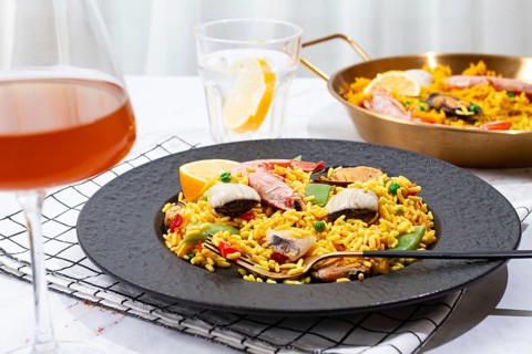 A table is set with a large black plate of seafood paella next to a golden pan. A glass with lemon