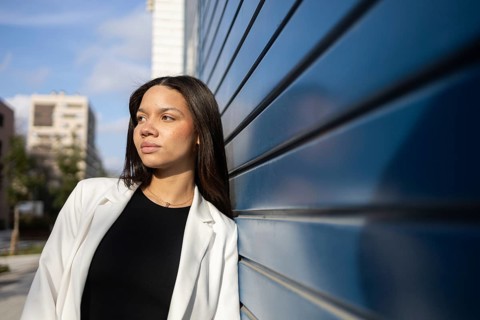 A confident woman in a white blazer leans against a blue wall, exuding professionalism in an urban