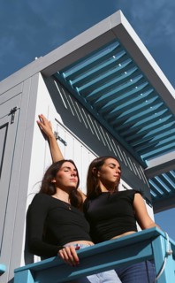 Two women friends enjoy a sunny day together, standing outdoors against a blue sky backdrop. They