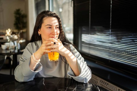 A mid-aged woman gazes out a window while sipping a drink in a sunlit, embodying a moment of