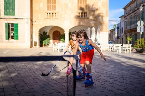 Two children play street hockey on roller skates in a sunlit plaza, showcasing teamwork and energy.