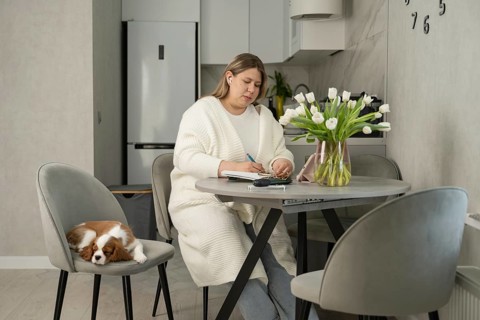 A woman in a cozy room writes at a round table while a small dog sleeps on a chair beside her. A