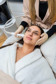 Woman lies on a bed in a robe while a beautician, wearing gloves, applies a radio frequency device