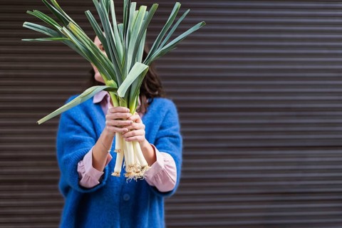 A woman in a blue sweater holds a bundle of fresh vegetables, hiding her face, against a blurred