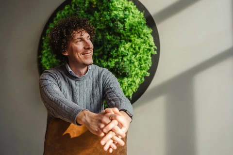 A young curly-haired man smiles while sitting in front of a vibrant green wall, symbolizing themes