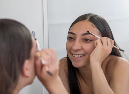 A woman smiles while using an eyebrow pencil in front of a mirror, focusing on perfecting her brows