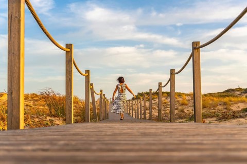 Woman in dress enjoying freedom and leisure on a wooden boardwalk, walking towards sandy dunes and