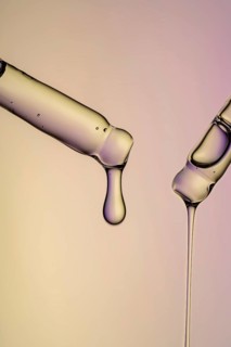 Close-up of face serum dripping from glass pipettes against a softly lit, colored background,