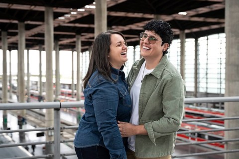 A joyful lesbian couple shares a moment of laughter in a train station. They are embracing each