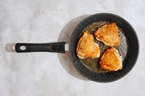 Three chicken thighs are sizzling in a frying pan on a marble countertop. The cooking process shows