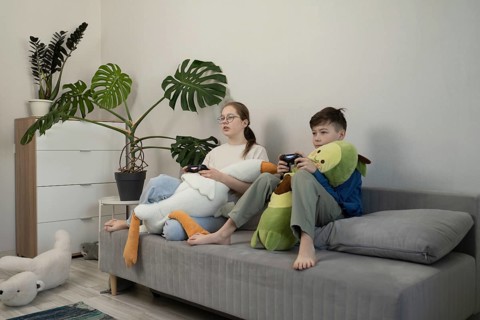 Two children sit on a gray couch, playing video games with controllers in hand. Plush toys rest on