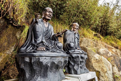 Two meditating monk statues sit on stone platforms against a lightly wooded background, The Buddha