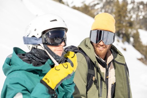 Two snowboarders equipped with gear enjoying a sunny day in the snowy backcountry. They are dressed