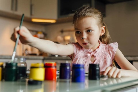 A young girl in a pink shirt focuses intently as she uses a brush to paint with colorful paints set
