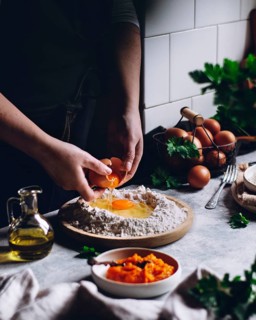 A person cracks an egg into a mound of flour, surrounded by a rustic kitchen setting with herbs,