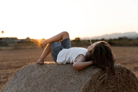 A child in casual summer attire lounges on a hay bale in a rural field as the sun sets, creating a