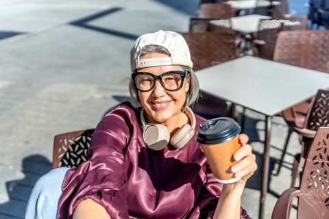 Woman with gray hair smiling at the camera, wearing a cap, glasses, and headphones around her neck,