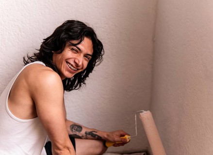 Cheerful young man with a brush in hand, painting a white wall in his home, smiling widely. His