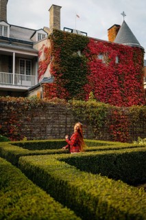 A woman in a red plaid coat stands in a manicured garden at a historic Montreal site, surrounded by