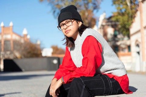 A young Latina in a vibrant red top and black beanie sits in a sunny urban setting Her casual