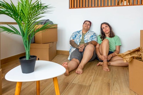 Lesbian friends sit on the floor amidst moving boxes, taking a break. A small table with a plant