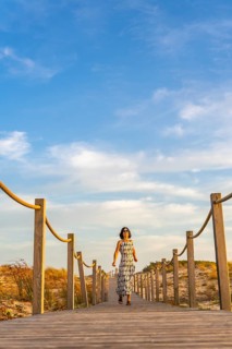 Woman in a flowing dress walking along a wooden boardwalk through sand dunes at sunset in