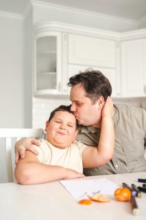 Father affectionately kisses his smiling son on the forehead in a cozy kitchen. Drawing supplies