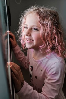 A young child with curly pink hair smiles while peering out a window. The child exudes a sense of