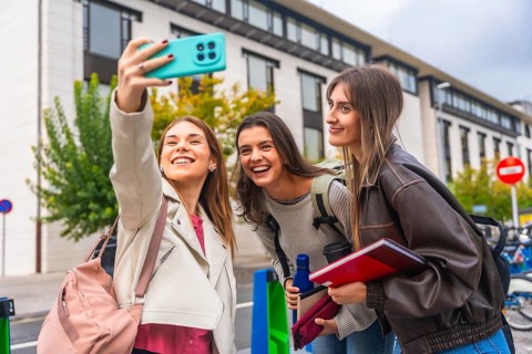 Three smiling young women students standing together outdoors on a university campus, taking a