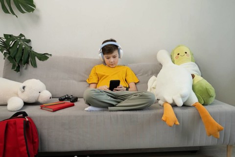 A child in headphones sits on a sofa, engrossed in a smartphone. Surrounded by plush toys and a