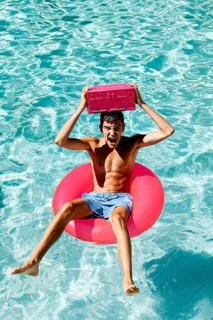 A joyful teenage boy is sitting on a pink float in a pool, holding a pink radio above his head. The