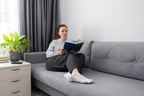 A woman sits comfortably on a gray sofa, reading a book. The serene atmosphere is enhanced by soft