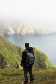 Traveler with backpack stands on a grassy cliff, gazing at a mist-covered fjord and mountains in