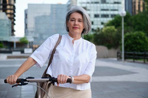 A mature woman with gray hair rides a scooter confidently in an urban environment. She wears a