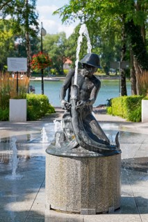 Fountain with statue in a sunny park with trees and water in the background, Ostroda, Osterode in
