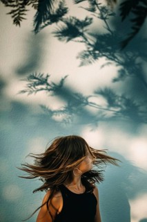 A teen girl stands in a sunlit area, her hair swirling gracefully. Tree shadows create an artistic