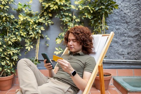A young man lounging outdoors in a chair, holding a smartphone and a credit card. He is casually