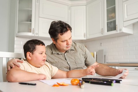A father helps his son with homework at the kitchen table. They are surrounded by school supplies,