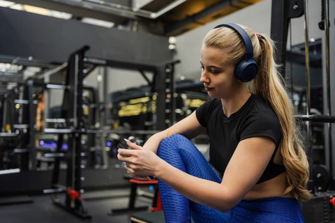 A blonde woman with headphones relaxes while checking her phone at a gym. She wears fitness attire