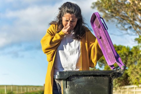 A woman in a yellow cardigan holds her nose while opening a trash bin in her backyard, representing