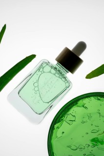 A bottle of aloe vera serum with a dropper lies on a white background, surrounded by green leaves.