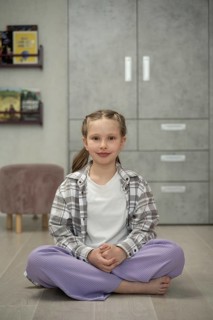 A young girl sits calmly on the floor with her legs crossed, smiling gently in a cozy, modern home