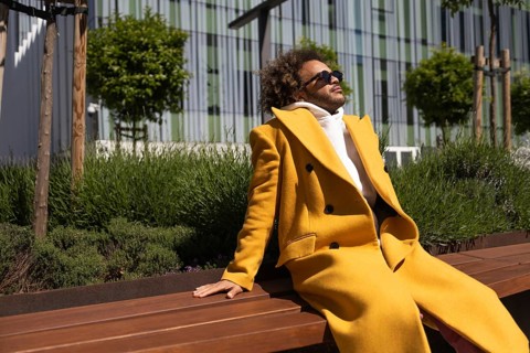Afro man relaxing on a wooden bench outdoors, wearing a fashionable yellow coat and sunglasses,