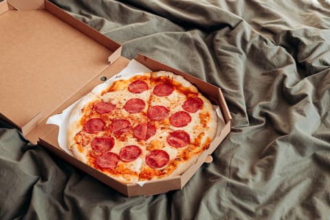 A freshly baked pepperoni pizza sits inside an open cardboard box placed on a crinkled gray bed