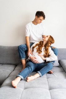 A young couple, expecting a baby, sits on a grey sofa at home. The woman gently cradles her rounded