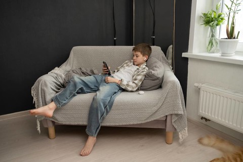 Child lounging on a sofa, using a smartphone with ear buds, in a cozy living room setting. Modern