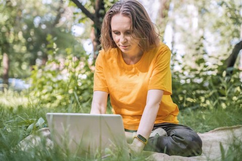 A woman sitting on a blanket in a lush park uses a laptop for remote work. Surrounding greenery and
