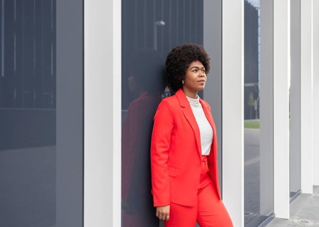 A woman in a vibrant red suit leans confidently against a modern building. Her thoughtful