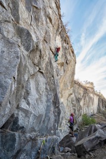 Two climbers face a steep rock face. One ascends the cliff while his partner secures the rope from