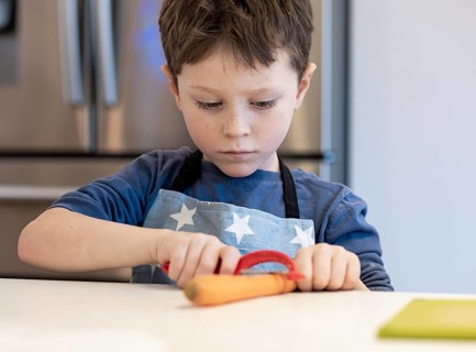 A focused young child wearing a star patterned apron peels a carrot in a bright kitchen environment